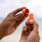 Bálsamo Labial Sun doTERRA SPF15 — mãos a abrir frasco na praia com fundo de mar, proteção solar natural