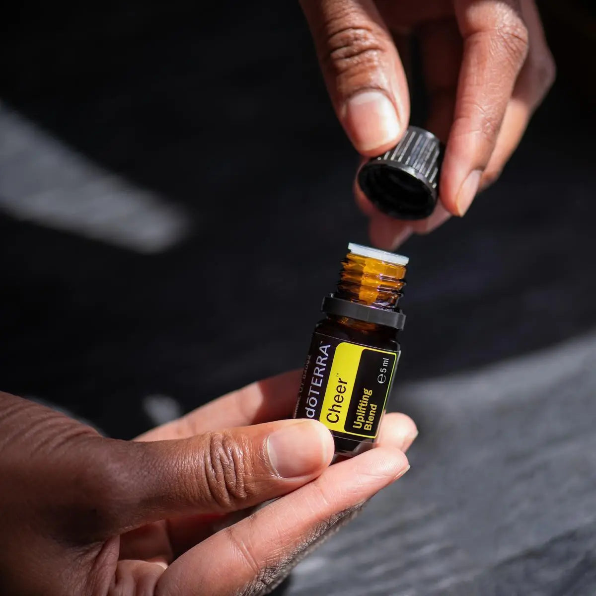 Cheer doTERRA 5ml duas mãos abrindo frasco fundo escuro dramático, alegria aromaterapia CPTG