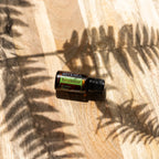 TerraShield doTERRA 15ml — tábua de madeira clara com sombras de fetos, vista zenital tropical, óleo essencial protetor