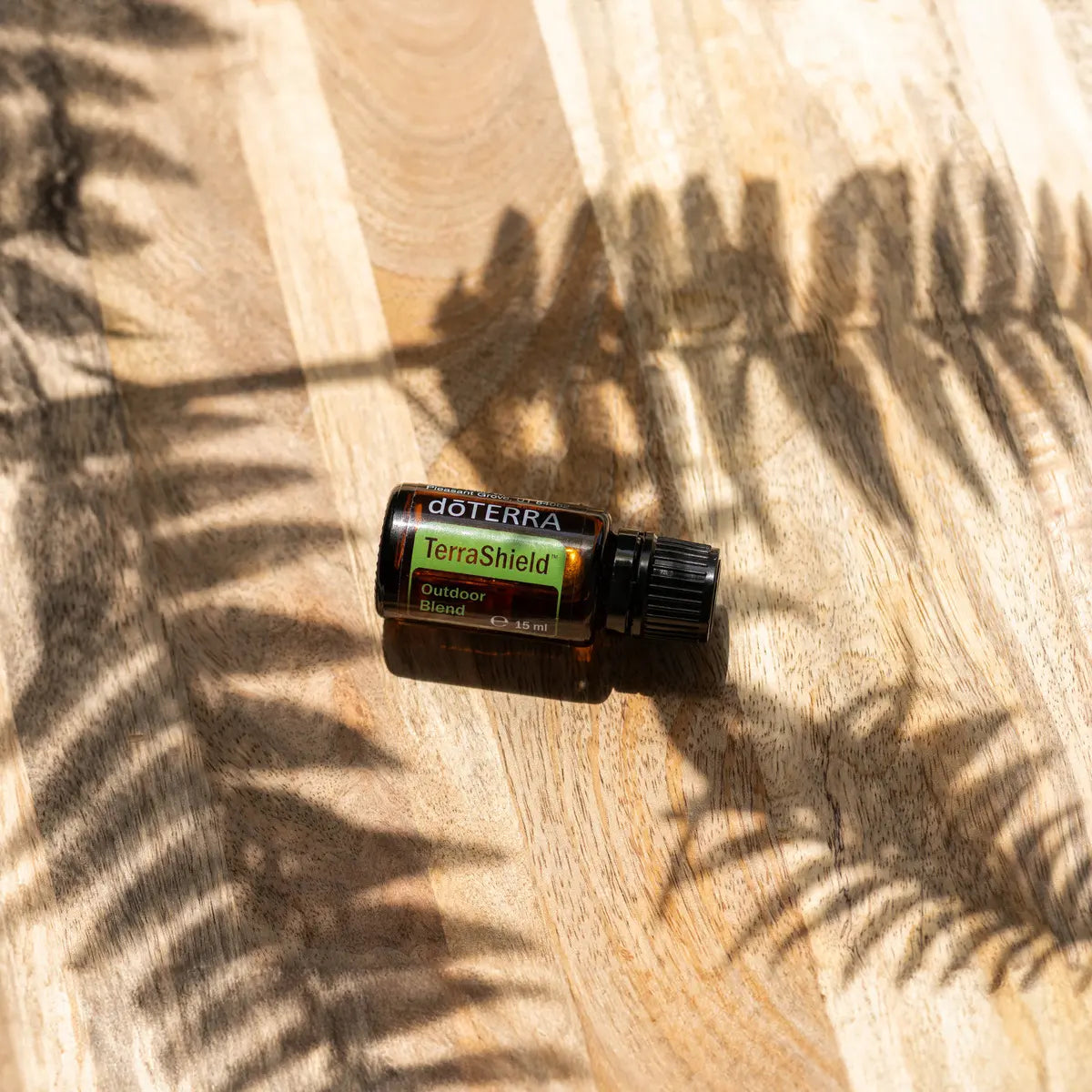 TerraShield doTERRA 15ml — tábua de madeira clara com sombras de fetos, vista zenital tropical, óleo essencial protetor