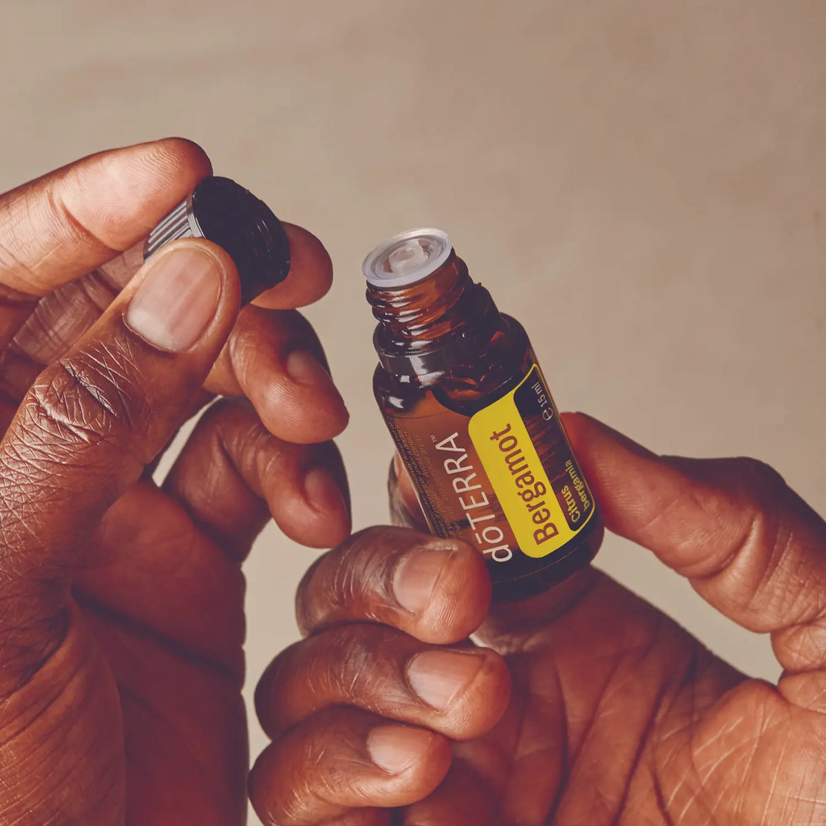 Óleo Essencial Bergamota doTERRA 15ml — mãos a abrir frasco com fundo bege, óleo essencial CPTG puro