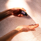 Óleo Essencial Patchouli doTERRA 15ml — mãos em palma seguram frasco com feixe de luz solar e sombras em parede rosa, aromaterapia, óleo essencial puro CPTG