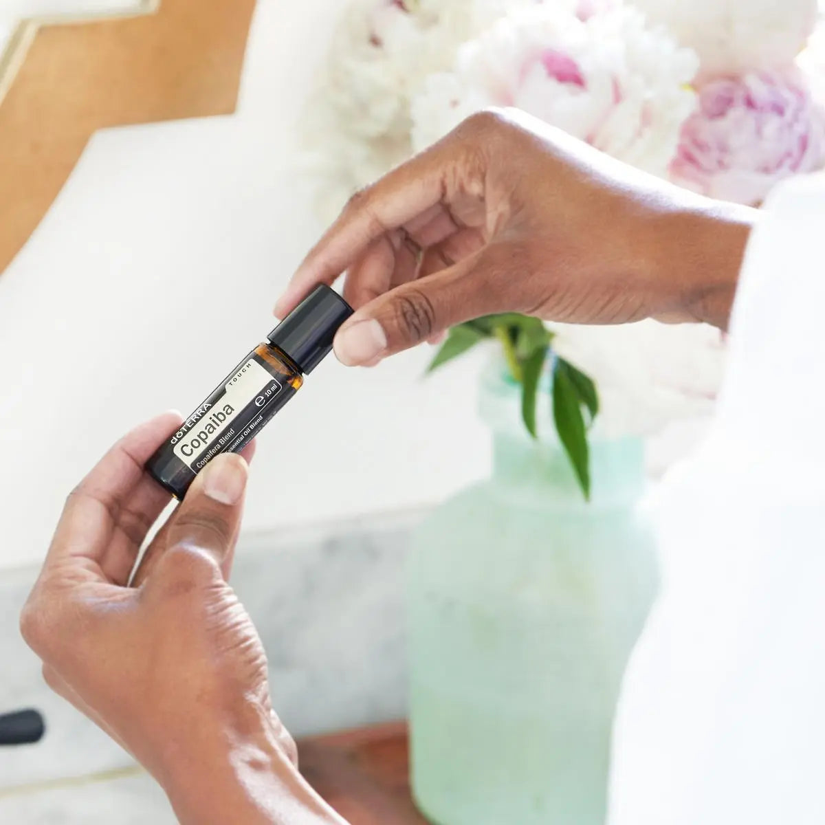 Copaiba Touch doTERRA Roll-on 10ml — mãos escuras com peonías rosas, óleo essencial calmante puro CPTG