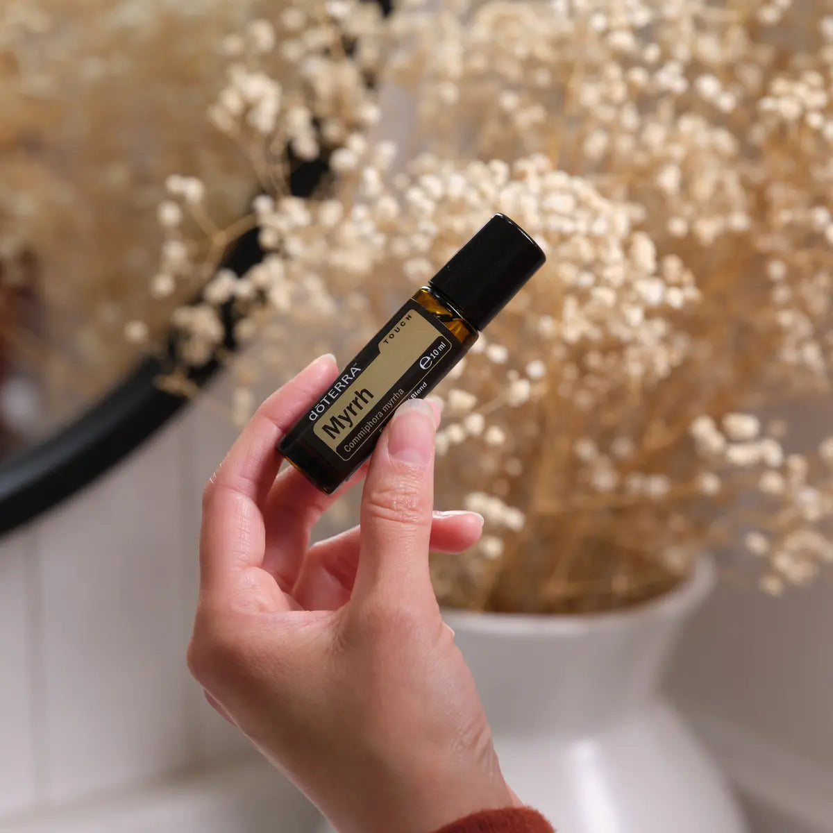 Roll-On Mirra Touch doTERRA 10ml — mão com flores secas gypsophila fundo difuminado, óleo essencial mirra puro CPTG