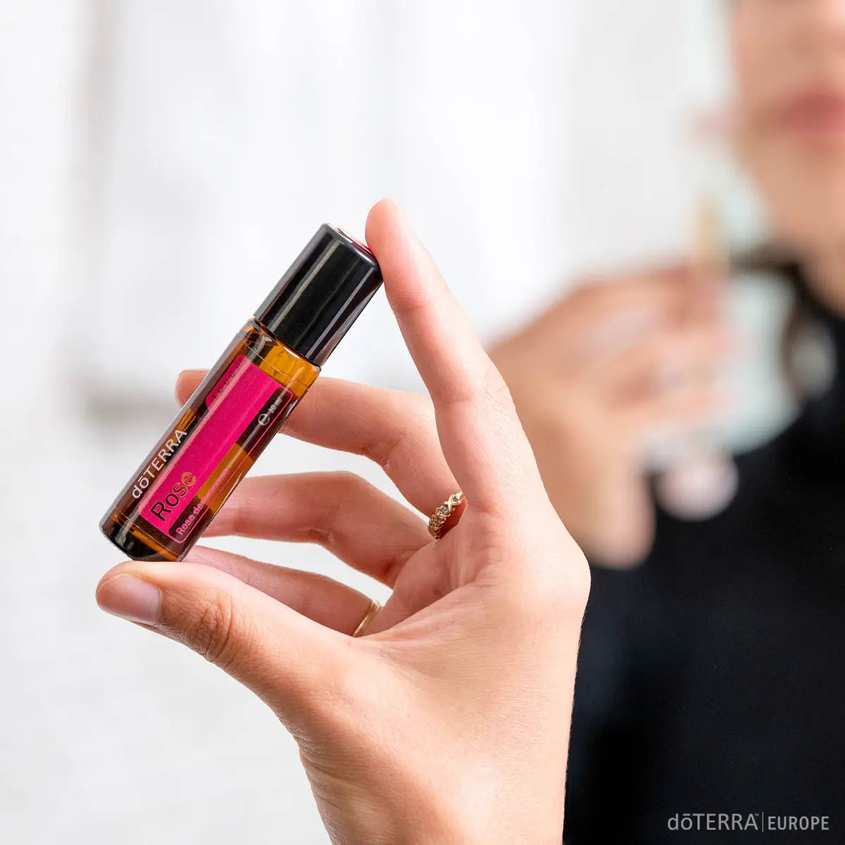 Roll-On Rosa doTERRA 10ml — mão de mulher a segurar frasco em interior com bokeh, óleo essencial rosa puro