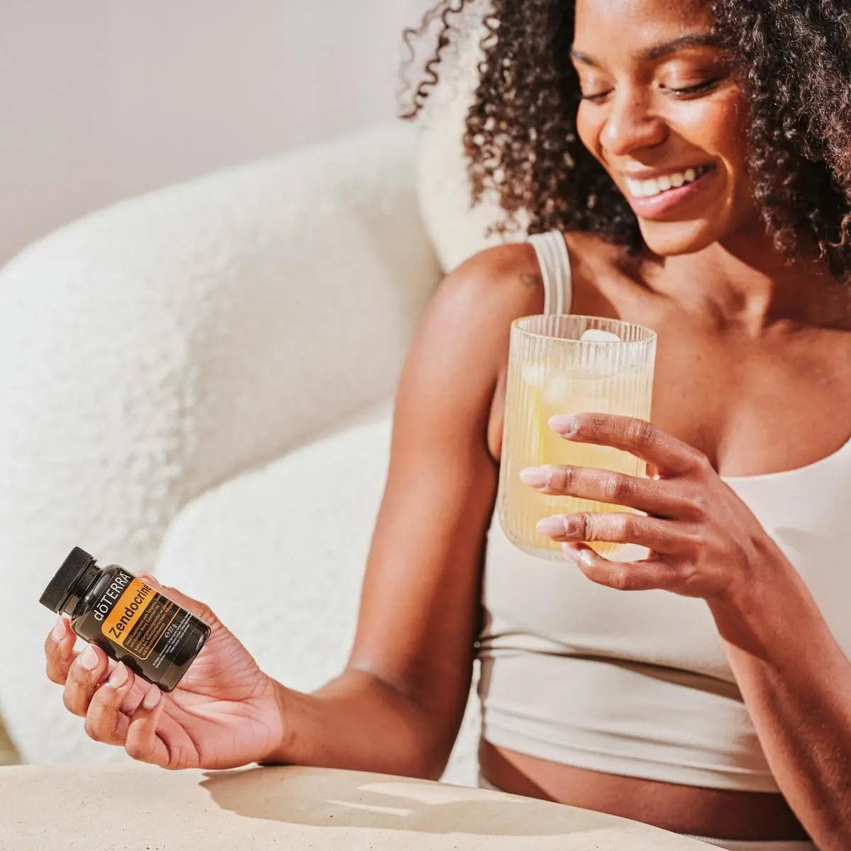 Cápsulas moles Zendocrine doTERRA mulher com cabelo encaracolado segurando frasco preto com vaso no sofá — suplemento detox natural CPTG